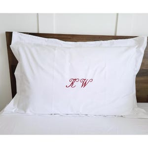 May include: White pillowcase with a red embroidered "KW" monogram. The rectangular pillowcase has a folded edge. It is placed on a bed with a wooden headboard. The bedding is white.