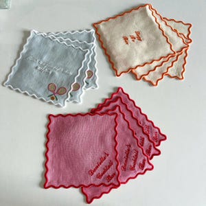 May include: A collection of square, scalloped-edge coasters in various colors. One set is light blue with tennis racket embroidery and text. Another is off-white with orange trim and text. The last set is pink with red trim and text.