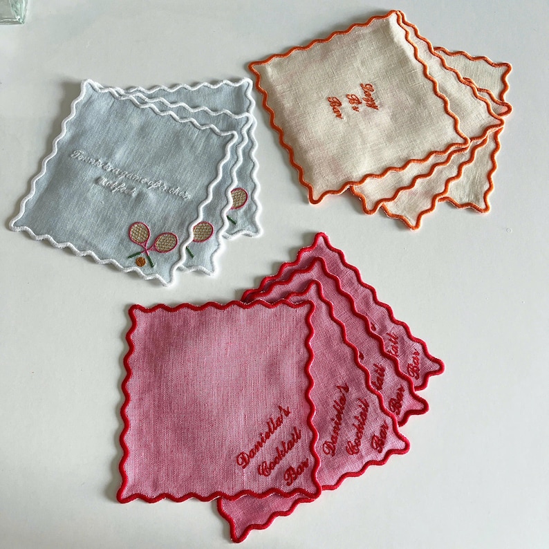 May include: A collection of square, scalloped-edge coasters in various colors. One set is light blue with tennis racket embroidery and text. Another is off-white with orange trim and text. The last set is pink with red trim and text.