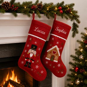 Könnte beinhalten: Zwei rote Weihnachtsstrümpfe mit weißem Wellensaum und gestickten Schneeflocken. Ein Strumpf zeigt einen Nussknacker und den Namen Lucas, der andere ein Lebkuchenhaus und den Namen Sophia in weißer Schreibschrift.