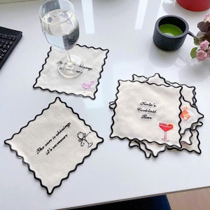 Op de afbeelding: Witte linnen cocktail onderzetters met zwarte geschulpte randen. Elke onderzetter heeft geborduurde tekst en een cocktail-thema illustratie. Eén onderzetter leest "The sun is shining it's summer". Een andere zegt "Kate's Cocktail Bar".