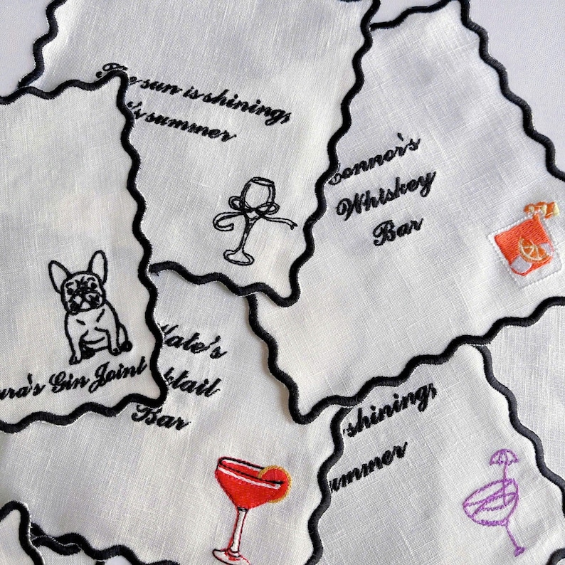 May include: White linen cocktail napkins with black scalloped edges. Each napkin features embroidered cocktail-themed designs and text, including a bulldog, a wine glass, and the words "Connor's Whiskey Bar."