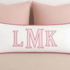 Op de afbeelding: Een wit rechthoekig kussen met roze rand en de geborduurde letters "LMK" in roze. Twee effen roze kussens staan op de achtergrond. Het kussen ligt op een lichtroze ondergrond.