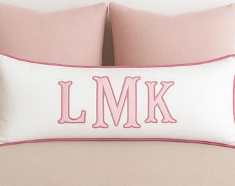 Może przedstawiać: Prostokątna biała poduszka lędźwiowa z różową lamówką na krawędziach i wyhaftowanymi różowymi literami monogramu LMK na środku. Poduszka znajduje się przed dwiema gładkimi, jasnoróżowymi kwadratowymi poduszkami dekoracyjnymi.