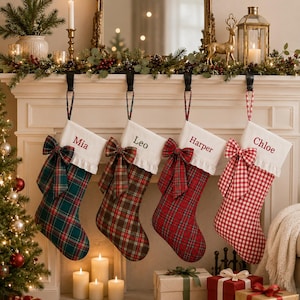 May include: Four Christmas stockings hanging in a row, each featuring a white cuff with a name embroidered in red or grey. The stockings display various plaid and gingham patterns in green, brown, and red, and each is adorned with a large fabric bow.