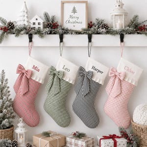 Puede incluir: Cuatro calcetines de Navidad personalizados colgados en fila. Cada calcetín tiene un puño blanco con un nombre bordado en rojo, verde, negro o rosa, y un lazo de tela grande. Los calcetines tienen rayas rojas, cuadros vichy verdes, rayas negras o cuadros vichy rosas.