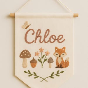 Puede incluir: Bandera crema bordada con el nombre "Chloe" en marrón. Presenta una mariposa, un zorro, setas, flores y una bellota. La bandera cuelga de un palito de madera con una cuerda.