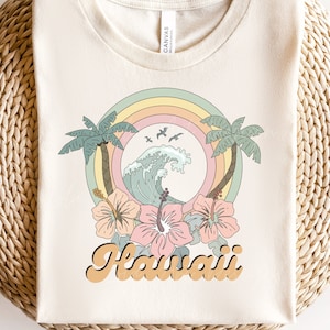 Könnte beinhalten: Ein weißes T-Shirt mit einem Retro-Design, das einen Regenbogen, Palmen, eine Welle und Hibiskusblüten zeigt. Der Text "Hawaii" ist in einer Retro-Schrift geschrieben.