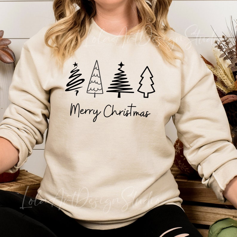 K&ouml;nnte beinhalten: Ein beiger Pullover mit einer schwarzen Grafik von vier stilisierten Weihnachtsb&auml;umen und dem Text "Frohe Weihnachten".