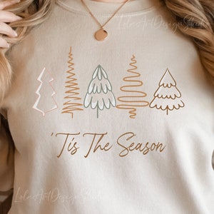 Könnte beinhalten: Ein beiger Pullover mit einem grafischen Design aus fünf stilisierten Weihnachtsbäumen in Braun-, Grün- und Weißtönen. Der Text "'Tis The Season" ist unter den Bäumen gedruckt.