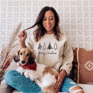 K&ouml;nnte beinhalten: Eine Frau tr&auml;gt einen hellbeigen Pullover mit einer schwarzen Grafik von stilisierten Weihnachtsb&auml;umen und dem Text "Merry Christmas". Sie sitzt auf einem Sofa mit einem braunen und wei&szlig;en Hund.