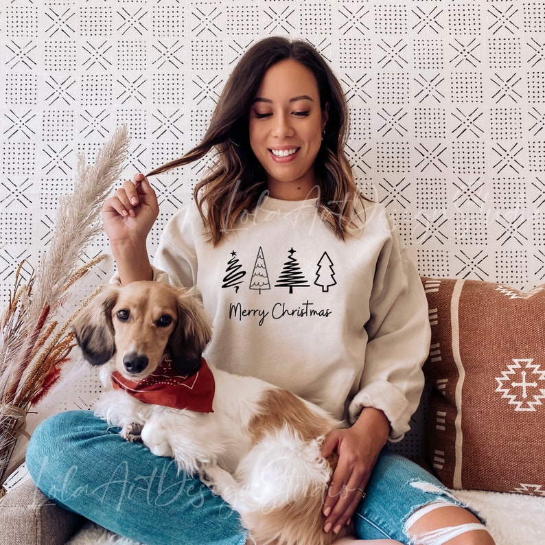 K&ouml;nnte beinhalten: Eine Frau tr&auml;gt einen hellbeigen Pullover mit einer schwarzen Grafik von stilisierten Weihnachtsb&auml;umen und dem Text "Merry Christmas". Sie sitzt auf einem Sofa mit einem braunen und wei&szlig;en Hund.