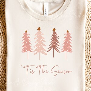 Könnte beinhalten: Ein weißes T-Shirt mit einer pinkfarbenen und braunen Grafik von vier stilisierten Weihnachtsbäumen mit goldenen Sternen oben. Der Text 'Tis The Season ist in Pink unter den Bäumen geschrieben.