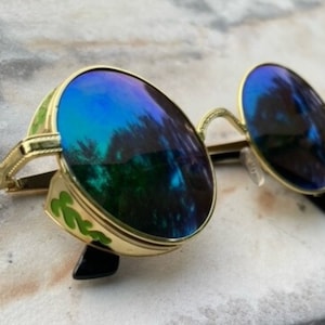 May include: Gold-framed sunglasses with round lenses reflecting a blue and green scene. The frames have a decorative green design on the sides and black temples. The lenses are a gradient of blue and green.