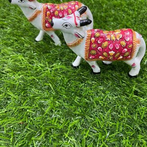 May include: Two white ceramic cow figurines with intricate painted details. The cows are decorated with pink, gold, and white floral patterns. The figurines are standing on a green artificial grass surface.