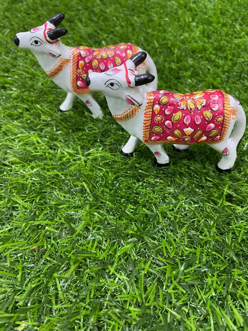 May include: Two white ceramic cow figurines with intricate painted details. The cows are decorated with pink, gold, and white floral patterns. The figurines are standing on a green artificial grass surface.