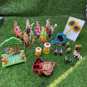 May include: A collection of colorful wooden figurines depicting a scene from Hindu mythology. The figurines are arranged on a green artificial grass surface. There are also several other decorative items, including a small wooden box, a mirror, and a peacock feather fan.
