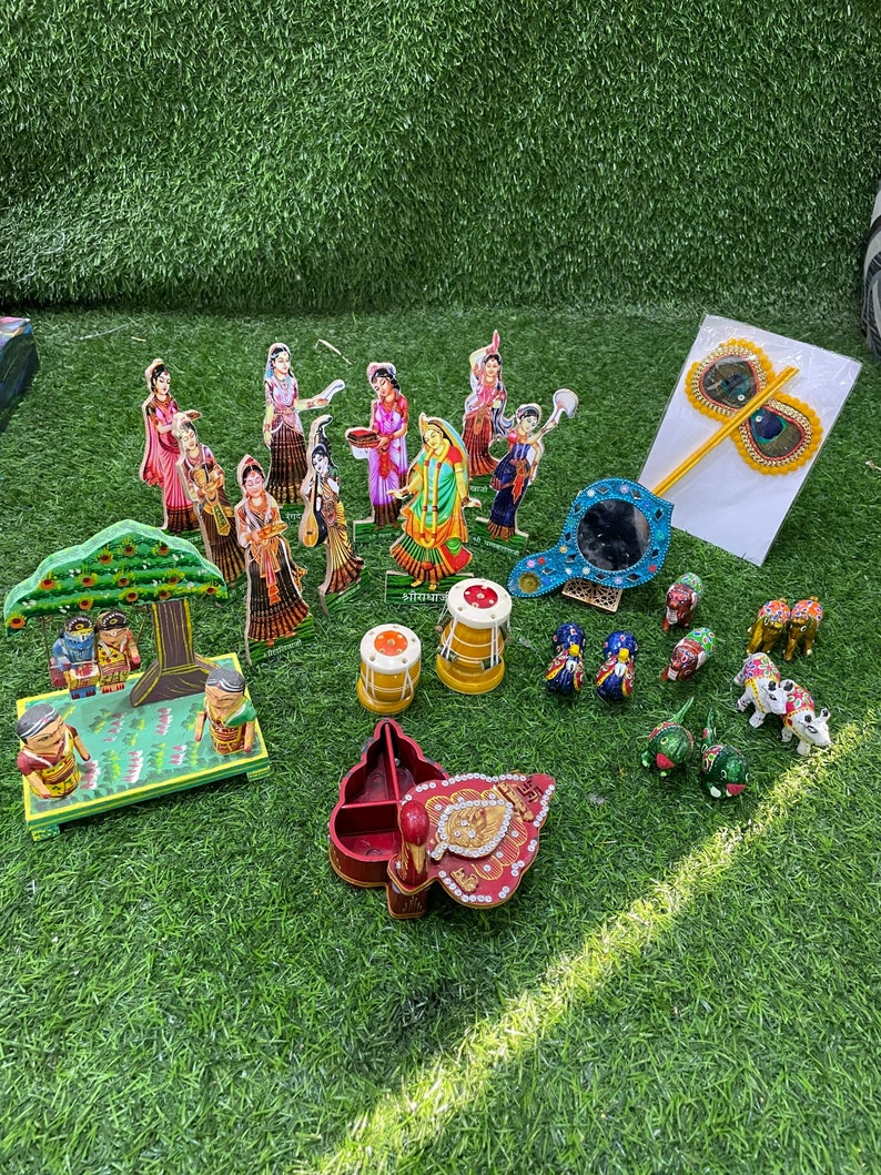 May include: A collection of colorful wooden figurines depicting a scene from Hindu mythology. The figurines are arranged on a green artificial grass surface. There are also several other decorative items, including a small wooden box, a mirror, and a peacock feather fan.