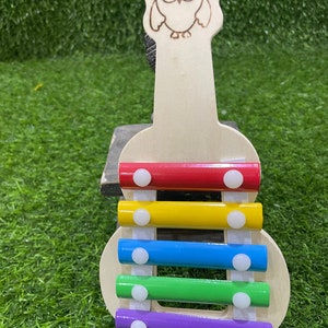 May include: A wooden xylophone shaped like a guitar with an owl design on the headstock. The xylophone has five bars in red, yellow, blue, green, and purple.