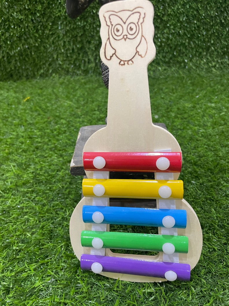 May include: A wooden xylophone shaped like a guitar with an owl design on the headstock. The xylophone has five bars in red, yellow, blue, green, and purple.