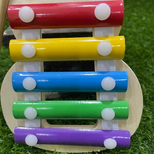 May include: A wooden xylophone toy with five colourful bars: red, yellow, blue, green, and purple. Each bar has two white plastic knobs on the ends.
