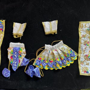 May include: A set of traditional Indian clothing for a doll, featuring a white skirt with colourful peacock designs, a purple top with white pearl accents, and a white sash with gold trim.