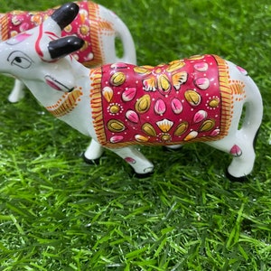 May include: Two white ceramic cows with intricate hand-painted details. The cows are decorated with pink, gold, and red patterns. The cows are standing on a green artificial grass surface.
