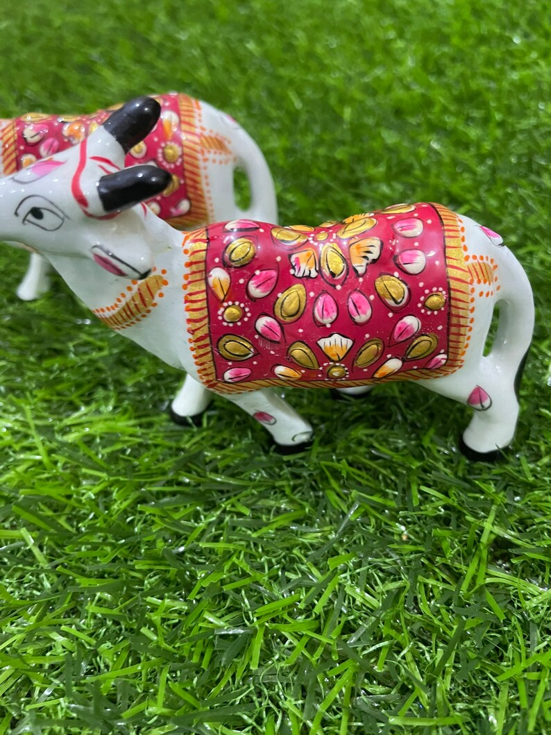 May include: Two white ceramic cows with intricate hand-painted details. The cows are decorated with pink, gold, and red patterns. The cows are standing on a green artificial grass surface.