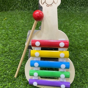 May include: A wooden xylophone shaped like an owl with a red mallet. The xylophone has five bars in rainbow colours: red, yellow, blue, green, and purple. Each bar has a white circle on top.
