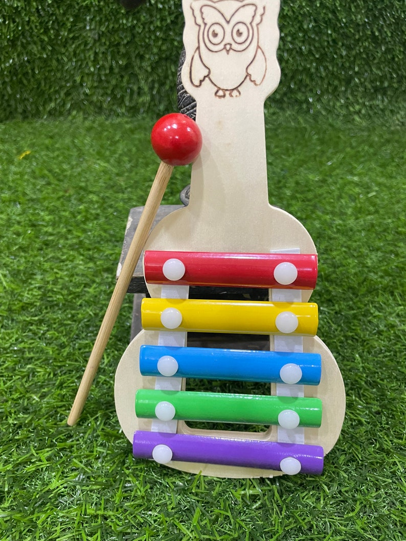 May include: A wooden xylophone shaped like an owl with a red mallet. The xylophone has five bars in rainbow colours: red, yellow, blue, green, and purple. Each bar has a white circle on top.