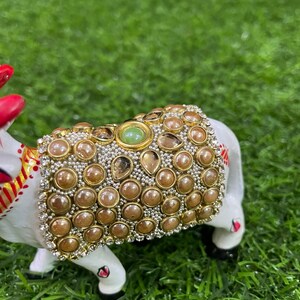 May include: A white ceramic figurine of a cow decorated with gold and brown beads and a green gemstone. The cow is standing on a green artificial grass surface.