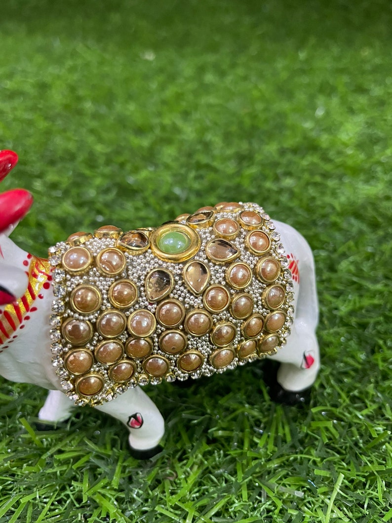 May include: A white ceramic figurine of a cow decorated with gold and brown beads and a green gemstone. The cow is standing on a green artificial grass surface.