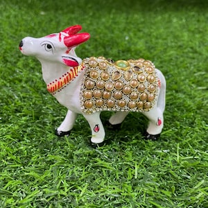 May include: White ceramic cow figurine with red horns and a decorative saddle adorned with gold and pearl-like beads. The cow is standing on a green artificial grass surface.