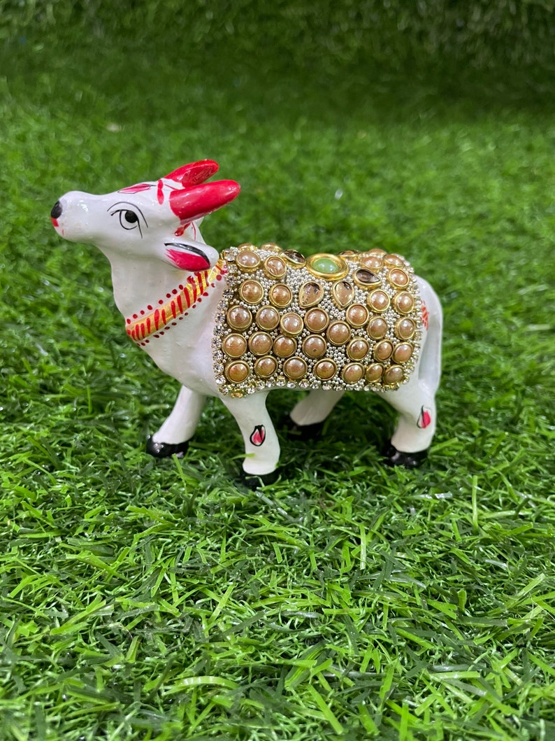 May include: White ceramic cow figurine with red horns and a decorative saddle adorned with gold and pearl-like beads. The cow is standing on a green artificial grass surface.