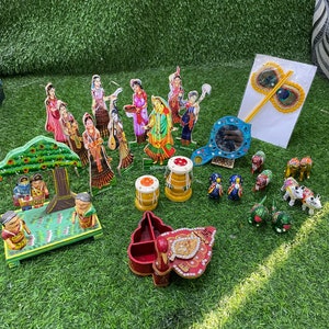 May include: A collection of colorful wooden figurines, including a peacock, cows, and a tree with two figures. There are also two small drums, a mirror, and a peacock feather wand. The items are arranged on a green artificial grass surface.