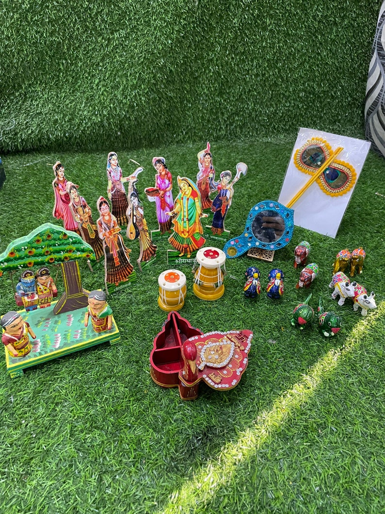 May include: A collection of colorful wooden figurines, including a peacock, cows, and a tree with two figures. There are also two small drums, a mirror, and a peacock feather wand. The items are arranged on a green artificial grass surface.