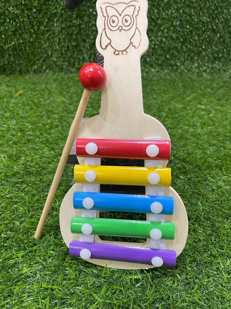 May include: A wooden xylophone shaped like an owl with a red mallet. The xylophone has five bars in rainbow colours: red, yellow, blue, green, and purple.