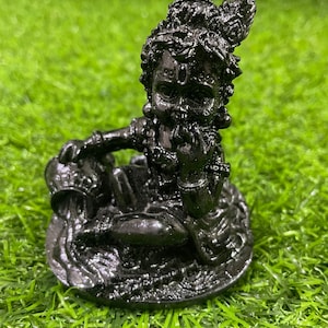 Puede incluir: Una figura negra de una deidad sentada, posiblemente Krishna, con detalles intrincados. La figura está adornada con joyas y una corona, sosteniendo una jarra. La estatua está colocada sobre una base redonda, sobre un fondo de césped artificial verde.