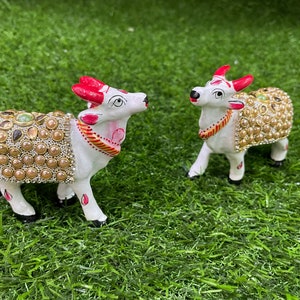May include: Two white ceramic figurines of cows decorated with gold and pearl-like embellishments. The cows are standing on a green artificial grass surface.