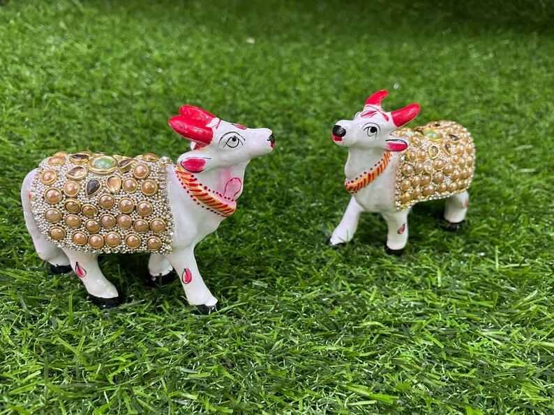 May include: Two white ceramic figurines of cows decorated with gold and pearl-like embellishments. The cows are standing on a green artificial grass surface.