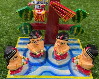 May include: A colorful wooden figurine depicting a scene from Hindu mythology. A blue figure with a crown sits atop a tree, while four figures in red and white clothing dance around a blue and white base.