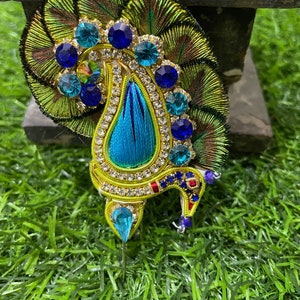 May include: A peacock feather design hair ornament with blue and green rhinestones and a blue and gold paisley design. The ornament is on a green background.