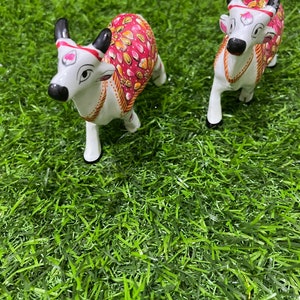 May include: Two white ceramic cow figurines with pink and gold painted designs. The cows are standing on a green artificial grass surface.