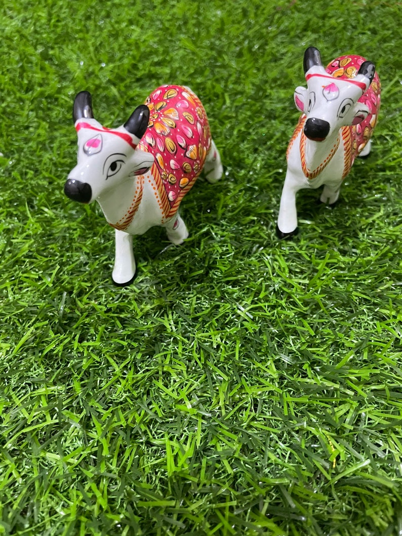 May include: Two white ceramic cow figurines with pink and gold painted designs. The cows are standing on a green artificial grass surface.