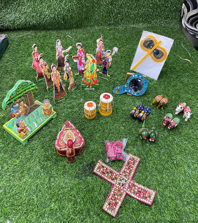 May include: A collection of colorful wooden figurines, including a group of people, a tree, and animals. There are also several decorative items, such as a mirror, a peacock feather, and a small box. The items are arranged on a green artificial grass surface.
