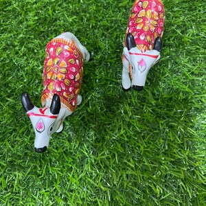 May include: Two white painted figurines of cows with red, pink, and gold floral designs on their backs. The cows are standing on a green artificial grass surface.