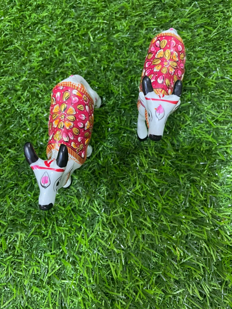 May include: Two white painted figurines of cows with red, pink, and gold floral designs on their backs. The cows are standing on a green artificial grass surface.