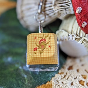 May include: A small, square pendant with a yellow gingham pattern and a floral design featuring mushrooms and leaves. The pendant is attached to a silver chain, with a red and white mushroom figurine in the background.