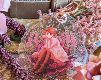 May include: A clear acrylic keychain featuring a seated figure in a red hat and dress, surrounded by pink flowers. The keychain has a gold-colored clasp. The background includes pink flowers and a decorative ribbon.