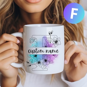 Può includere: Una tazza in ceramica bianca con un contorno nero della lettera "F" circondata da disegni di fiori in nero. I fiori sono su uno sfondo ad acquerello di viola, blu e rosa. Il testo "custom name" è stampato su uno striscione bianco nella parte inferiore della tazza.